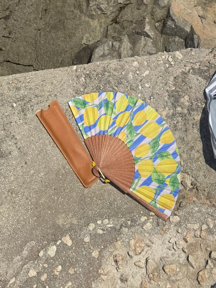 A striped gray and white beach towel and a colorful patterned folding fan with a brown handle rest on a concrete ledge overlooking a rocky coastline and turquoise ocean water.