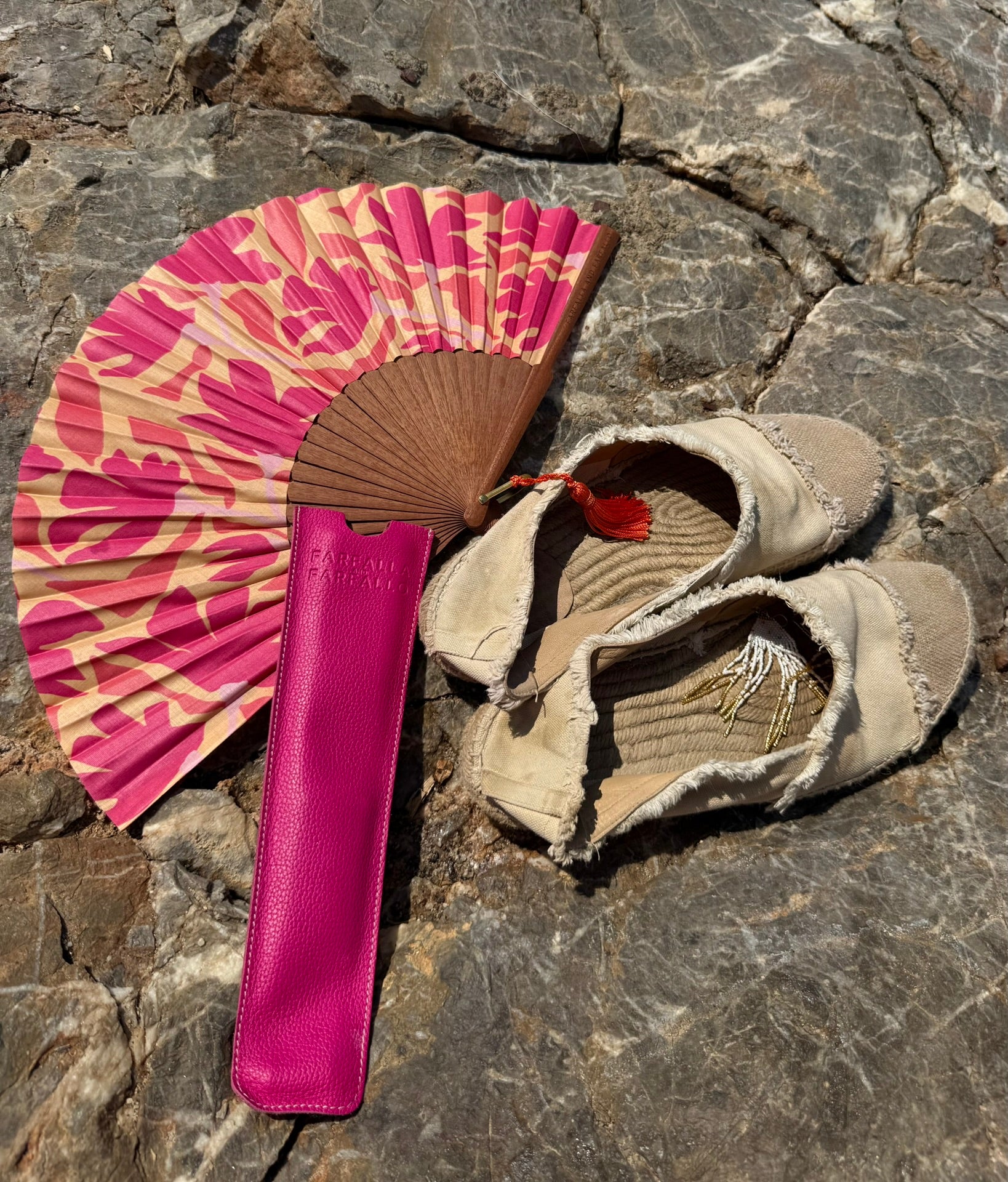 A pair of beige fringed leather sandals next to a pink and beige patterned folding fan and a matching pink leather case, arranged on a gray stone surface.