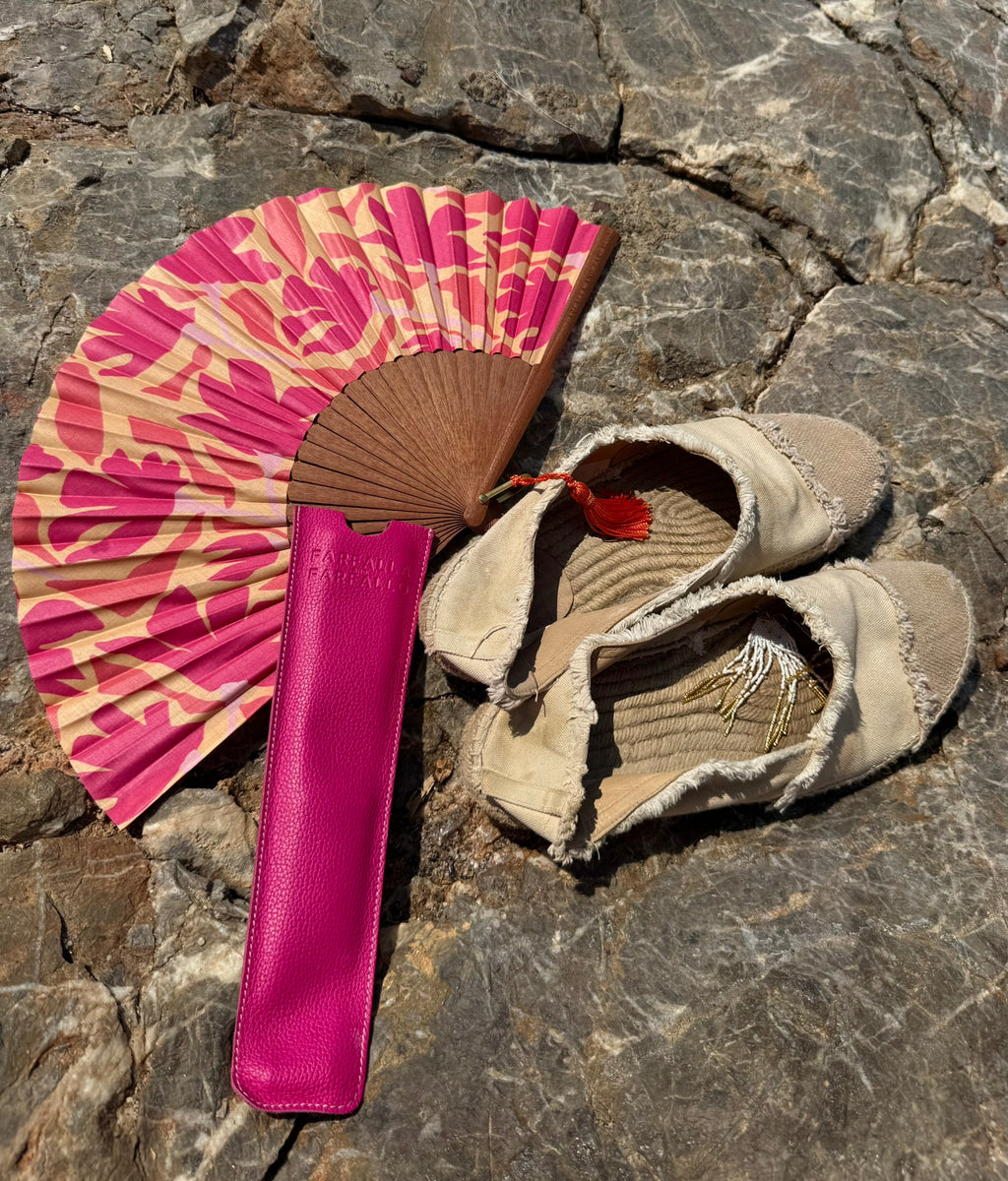 A pair of beige fringed leather sandals next to a pink and beige patterned folding fan and a matching pink leather case, arranged on a gray stone surface.