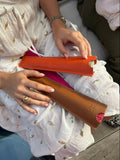 A person in a white embroidered dress holds two slim leather accessories, one orange and one brown, on their lap. The photo is cropped to show the hands, dress, and part of the seated setting.