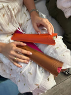 A person in a white embroidered dress holds two slim leather accessories, one orange and one brown, on their lap. The photo is cropped to show the hands, dress, and part of the seated setting.