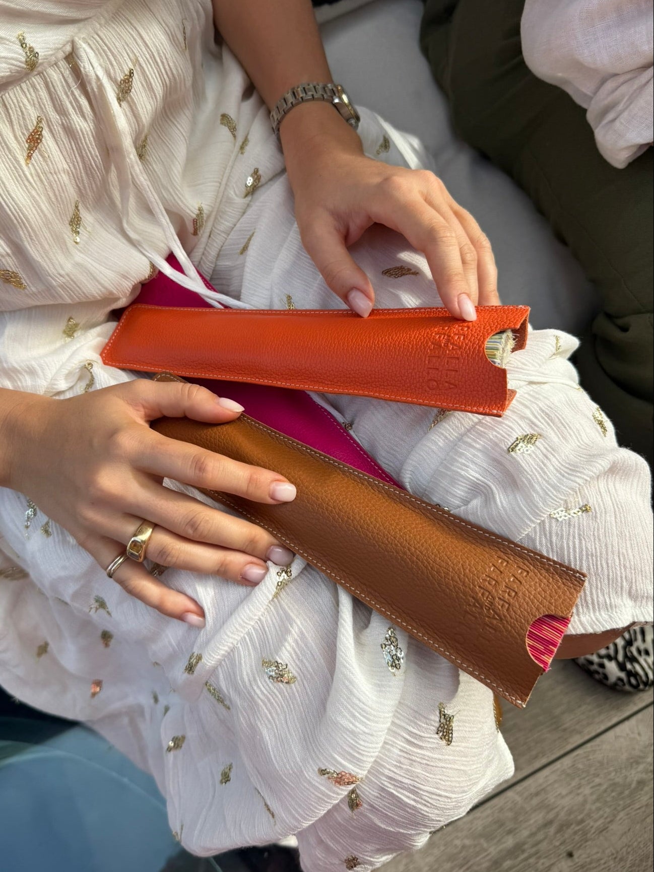 A person in a white embroidered dress holds two slim leather accessories, one orange and one brown, on their lap. The photo is cropped to show the hands, dress, and part of the seated setting.